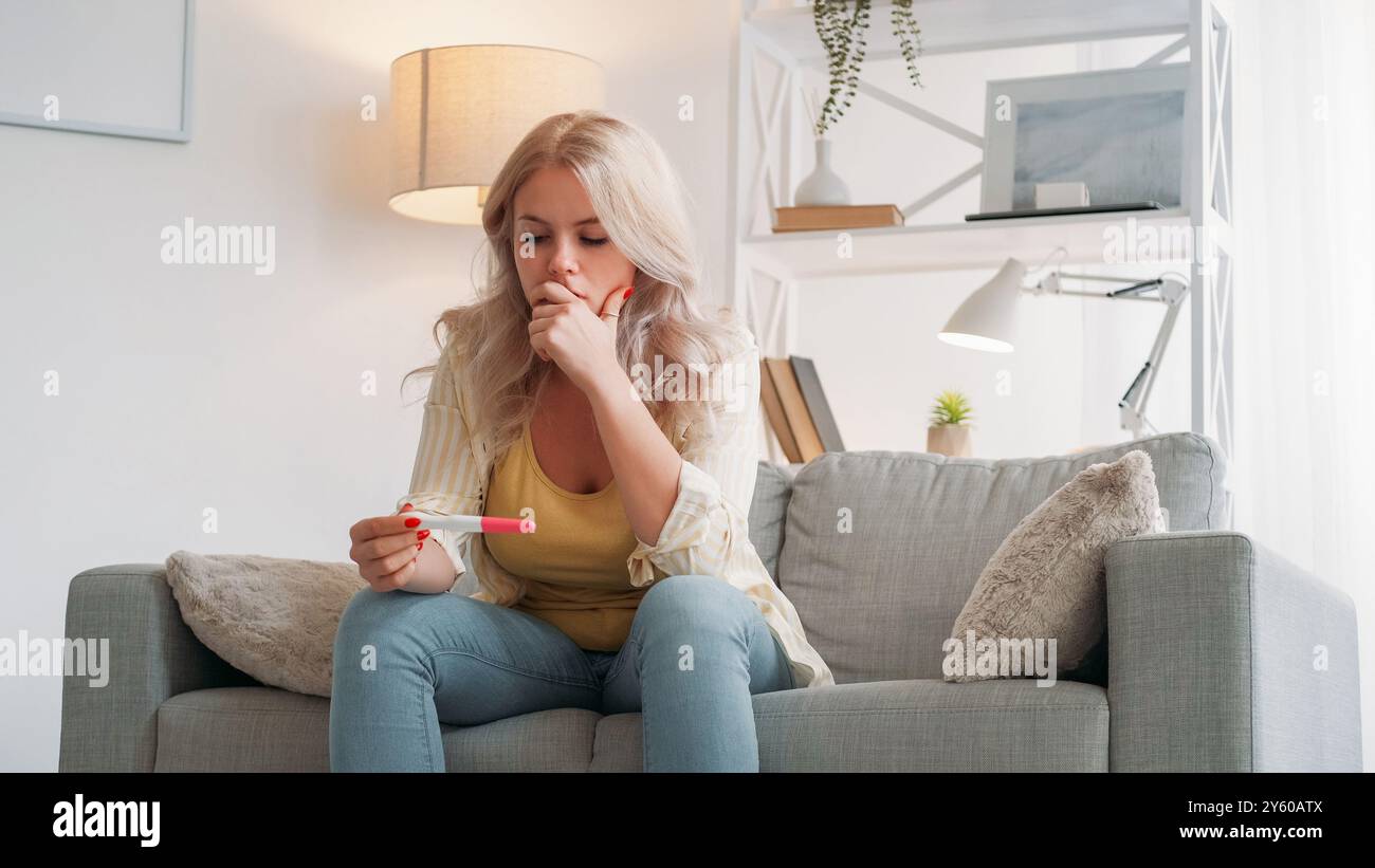 Grossesse non planifiée. Femme choquée. Regret de déception. Contrarié dame se sentant désespéré tenant le résultat du test assis canapé lumière intérieur de la pièce. Banque D'Images