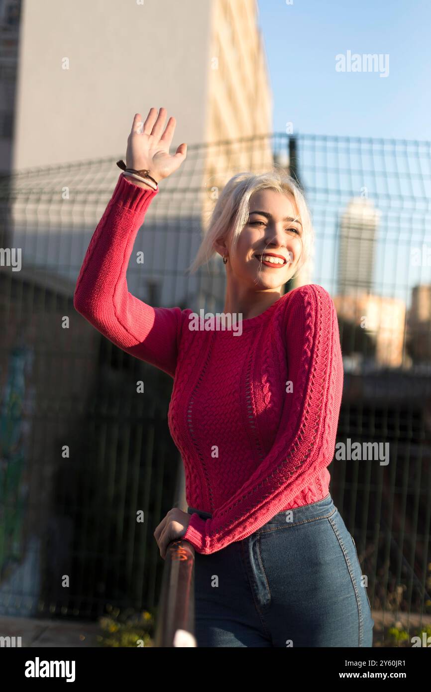 Une jeune femme joyeuse dans un pull rouge ondule avec un sourire éclatant, profitant du cadre urbain ensoleillé, ses cheveux blonds platine brillants dans la lumière. Banque D'Images