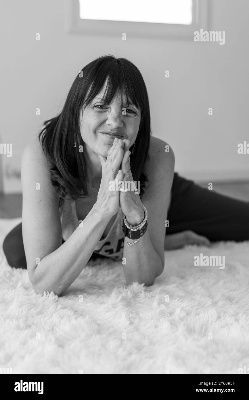 Portrait joyeux d'une femme mi-adulte reposant sur le tapis blanc moelleux dans son studio de yoga tout en souriant à la caméra. Banque D'Images