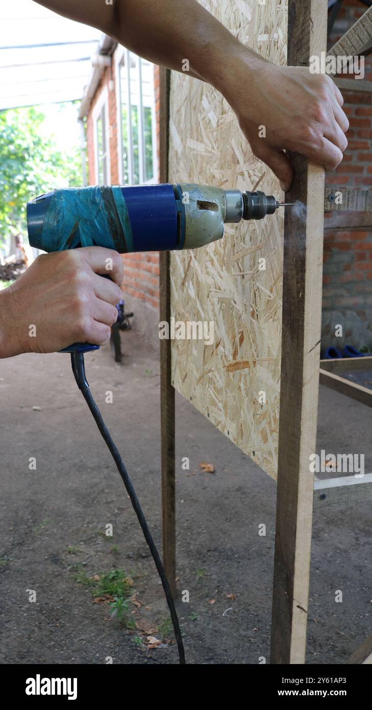 percer des trous dans la structure de cage de volaille avec la vieille perceuse dans les mains mâles, construire la cage de caille à l'aide de l'outil à main Banque D'Images
