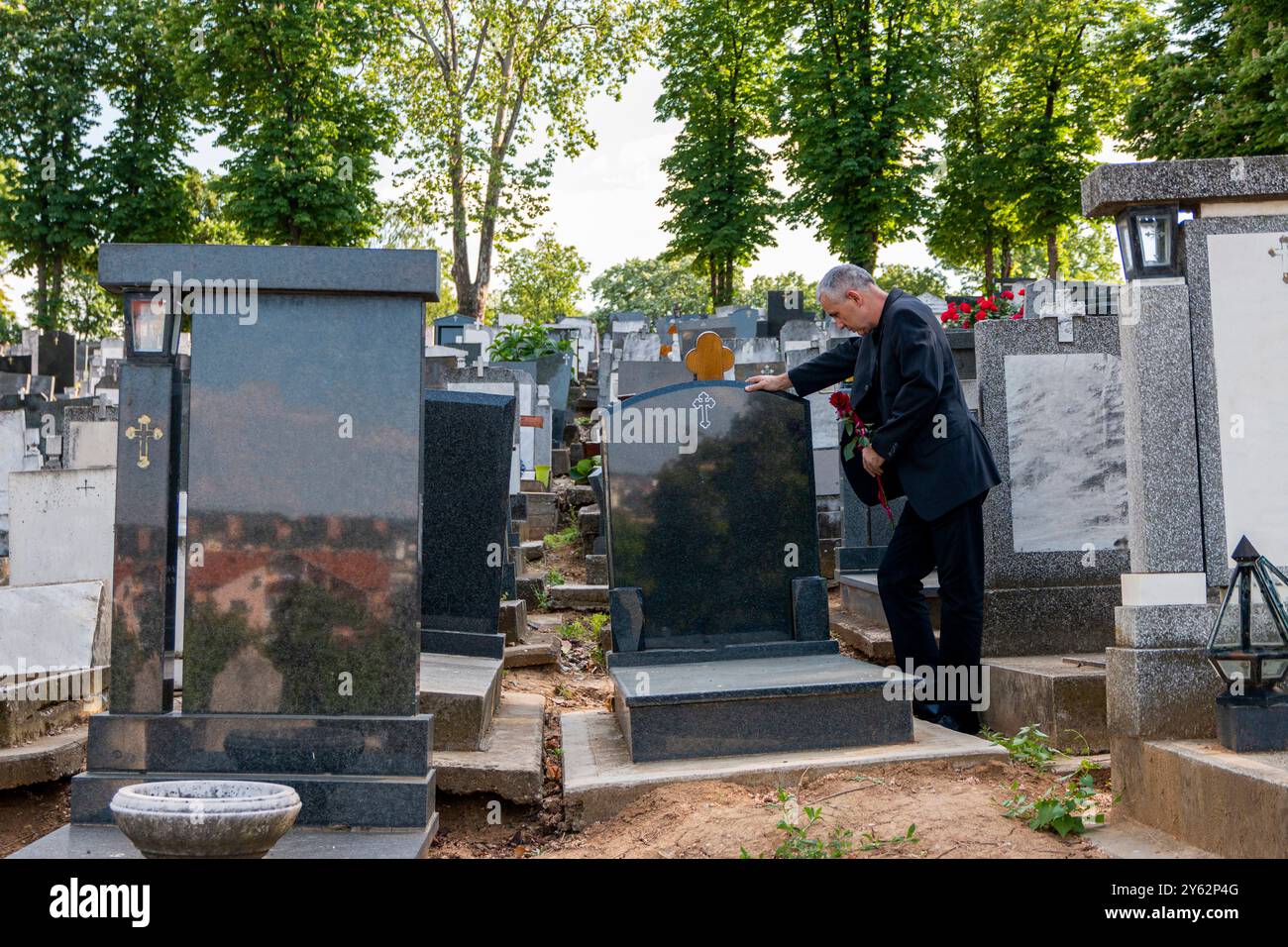 Homme mûr en vêtements noirs sur le cimetière, tenant une fleur et pleurant pour la perte de la famille. Concept de mort, deuil, funérailles et spiritualité. Banque D'Images