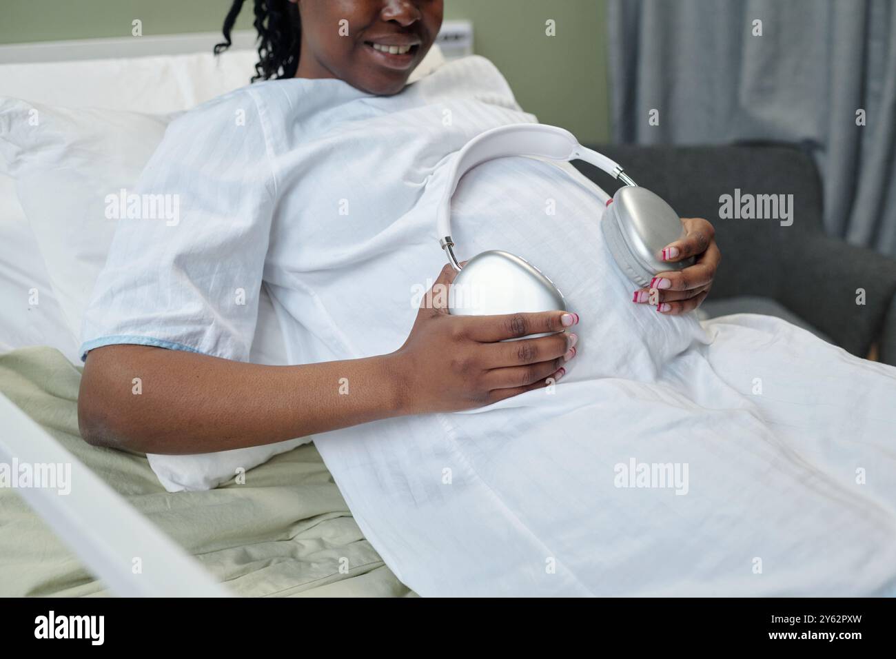 Femme enceinte assise dans une chambre d'hôpital utilisant un dispositif médical pour la surveillance. Elle sourit en portant une robe d'hôpital et assise sur le lit Banque D'Images