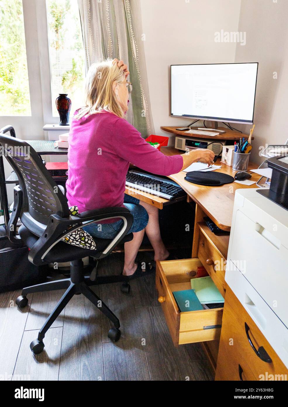 Une travailleuse à domicile, offrant des services de secrétariat et d'administration, portant des lunettes, communiquant avec les clients, à son ordinateur de bureau à domicile, dans son espace de travail Banque D'Images