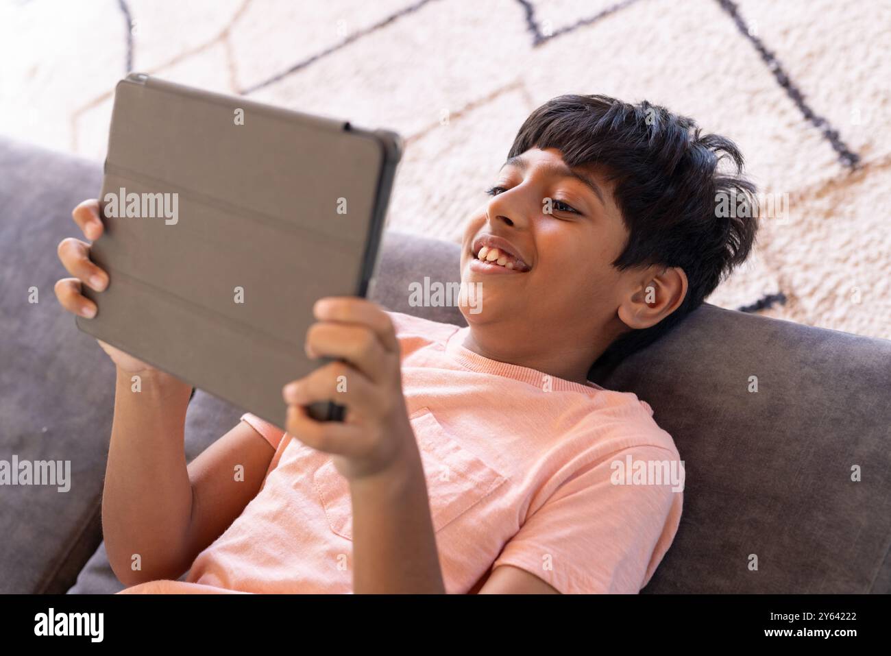 Garçon indien souriant à l'aide de la tablette tout en étant allongé sur le canapé à la maison, profitant du temps d'écran Banque D'Images