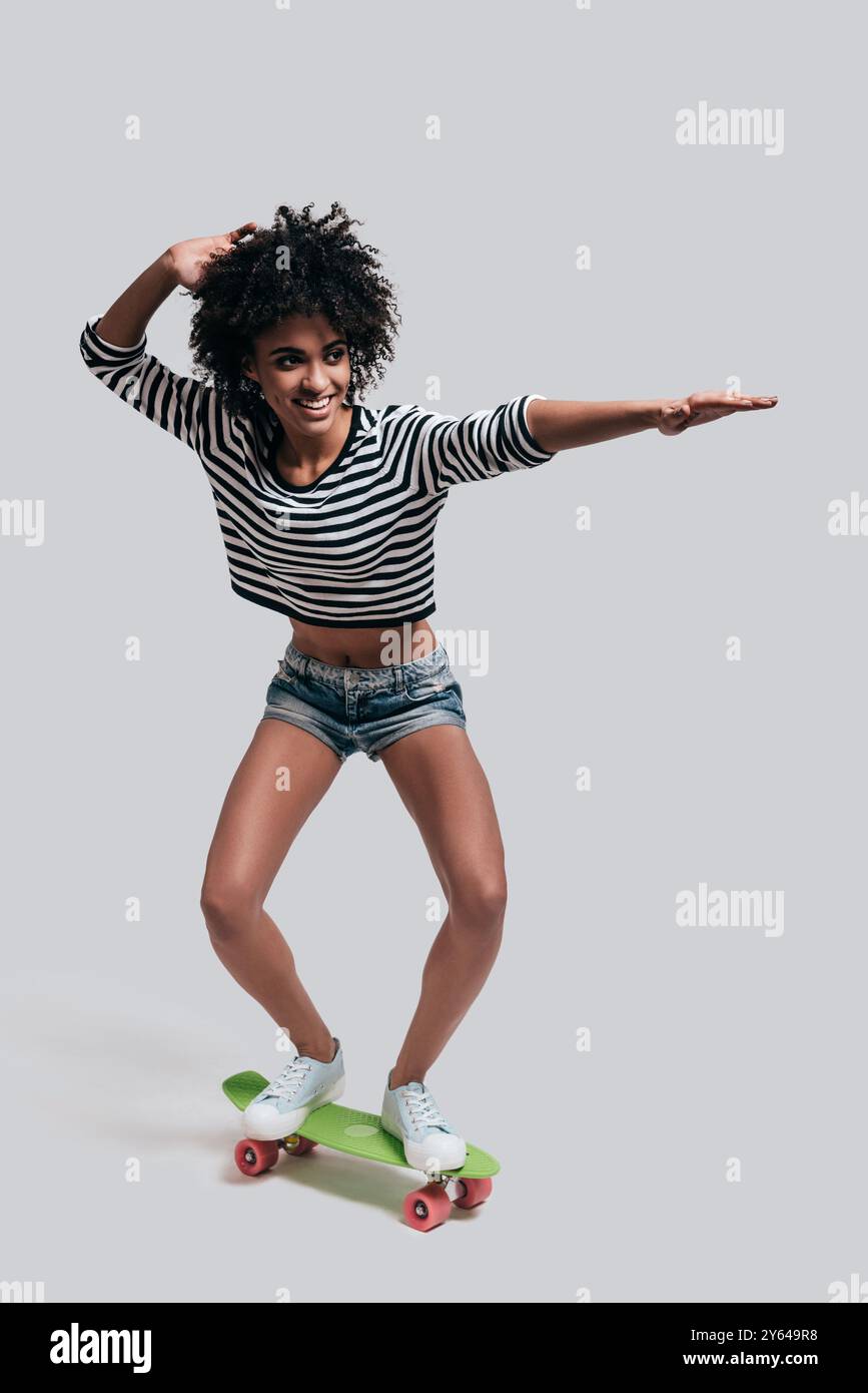J'aime le skateboard. Plein plan studio d'une jeune femme africaine ludique en tenue décontractée faisant des gestes et souriant tout en skateboard contre le gris Banque D'Images