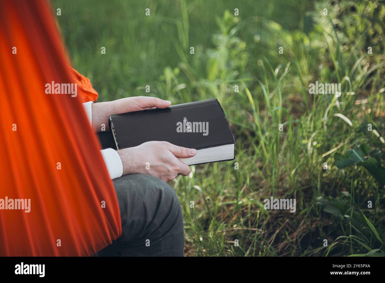 Vue rognée d'un individu chrétien engagé dans la lecture de la Sainte Bible tout en étant assis dans un hamac orange à l'extérieur. La verdure environnante s'améliore Banque D'Images