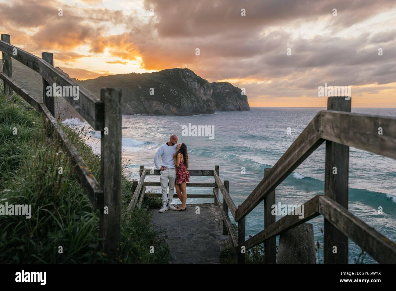 Un couple partage un moment tendre sur un escalier rustique en bois menant à une plage sereine, sur fond d'une falaise dramatique et d'un beau coucher de soleil Banque D'Images