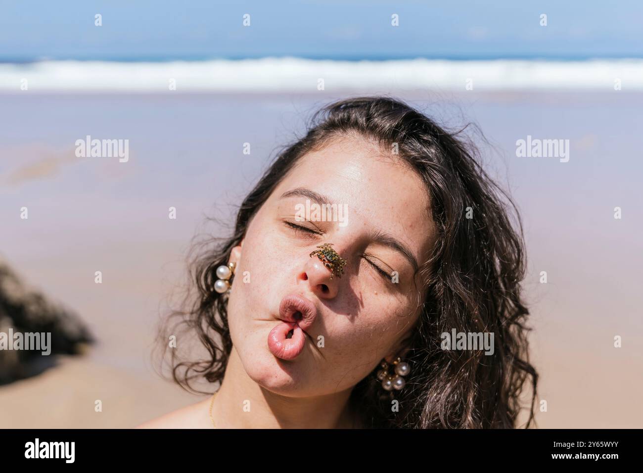 Une jeune femme ludique se fait un visage drôle avec un crabe sur son nez, un jour ensoleillé de plage, capturant un moment de joie et d'esprit insouciant Banque D'Images