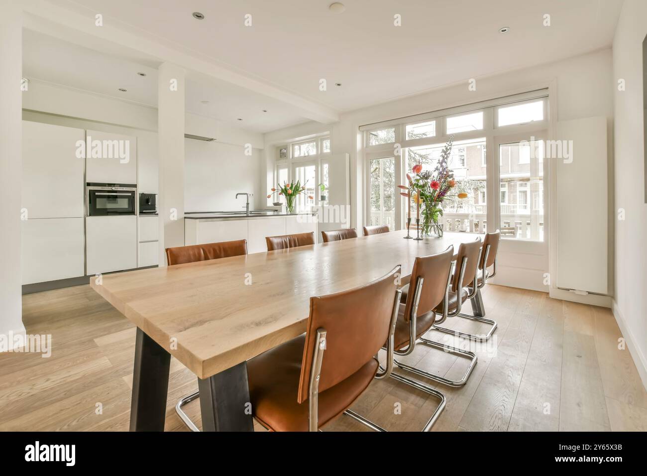 Une cuisine moderne spacieuse avec une grande table à manger, des chaises en cuir, des appareils encastrés et beaucoup de lumière naturelle provenant de grandes fenêtres. Banque D'Images