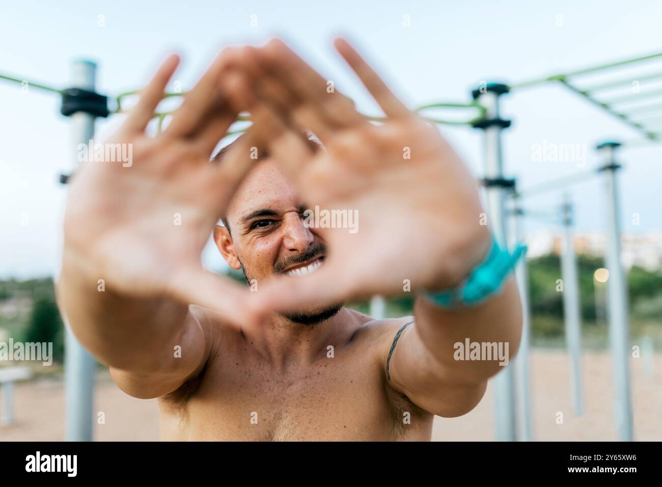 Un jeune homme en forme fait un cadre avec ses mains autour de la caméra dans un gymnase de calisthenics extérieur, mettant en vedette un moment de plaisir lors d'une sessio d'entraînement Banque D'Images