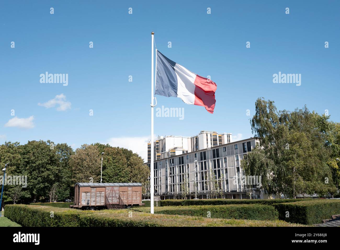 Le wagon commémoratif de la SNCF, installé en 1980, avec les bâtiments de la ville en arrière-plan, avec le drapeau français au centre. Le Mémorial de la Shoah à Drancy a été inauguré le 21 septembre 2012 par le président français François Hollande. Le mémorial se trouve en face de la Cité de la Muette et est une branche du Mémorial de la Shoah à Paris. France, Paris, 12 septembre 2024. La Muette s The cite, conçu dans les années 1930 pour fournir des logements à bas prix aux familles de Drancy, le complexe de bâtiments inachevés a servi de camp d'internement et plus tard de camp de concentration pour les Juifs. Près de 63 000 ont été expulsés. Banque D'Images