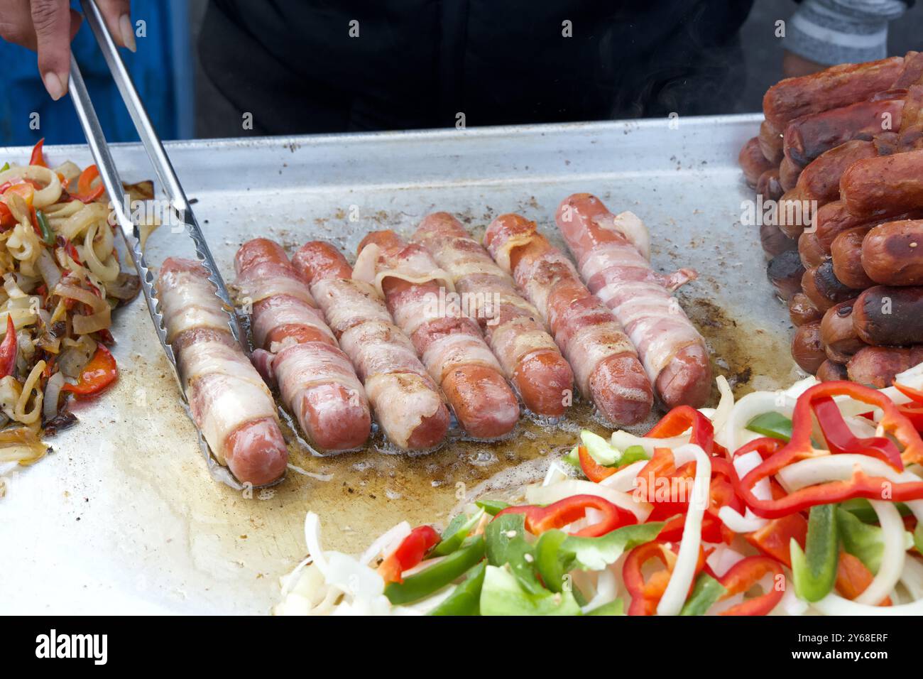 Vendeur de rue cuisant des hot dogs enveloppés de bacon, pinces tournant la nourriture. Cuisine populaire pour les foires et événements de rue Banque D'Images