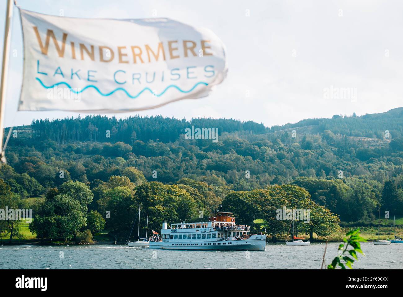 Un ferry navigue sur Windermere Lake Cruises dans le Lake District avec un drapeau qui souffle dans le vent, entouré de collines, d'arbres et d'eaux paisibles. Banque D'Images