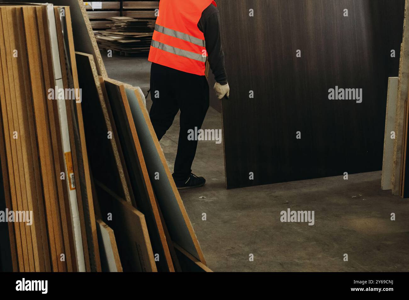 Comme la lumière du soleil filtre dans l'entrepôt, un travailleur diligent dans un gilet de sécurité lumineux transporte soigneusement une grande feuille de matériau au milieu de la stature imposante Banque D'Images