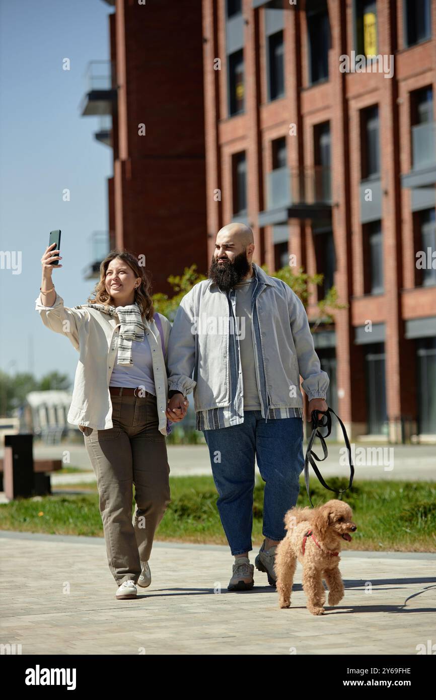 Couple heureux promenant chien tout en prenant selfie à l'extérieur près du complexe de construction moderne profitant d'une journée lumineuse et une atmosphère animée ensemble souriant tenant la main le long du chemin piétonnier Banque D'Images