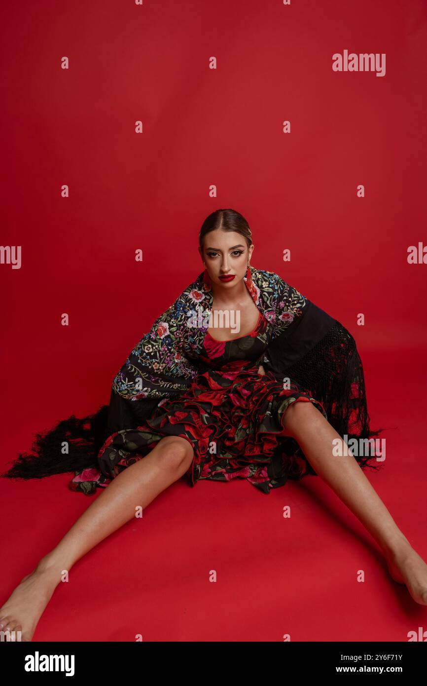 Une jeune femme dans une robe florale traditionnelle et châle est assise sur un fond rouge, mettant en valeur la mode et la culture. Banque D'Images