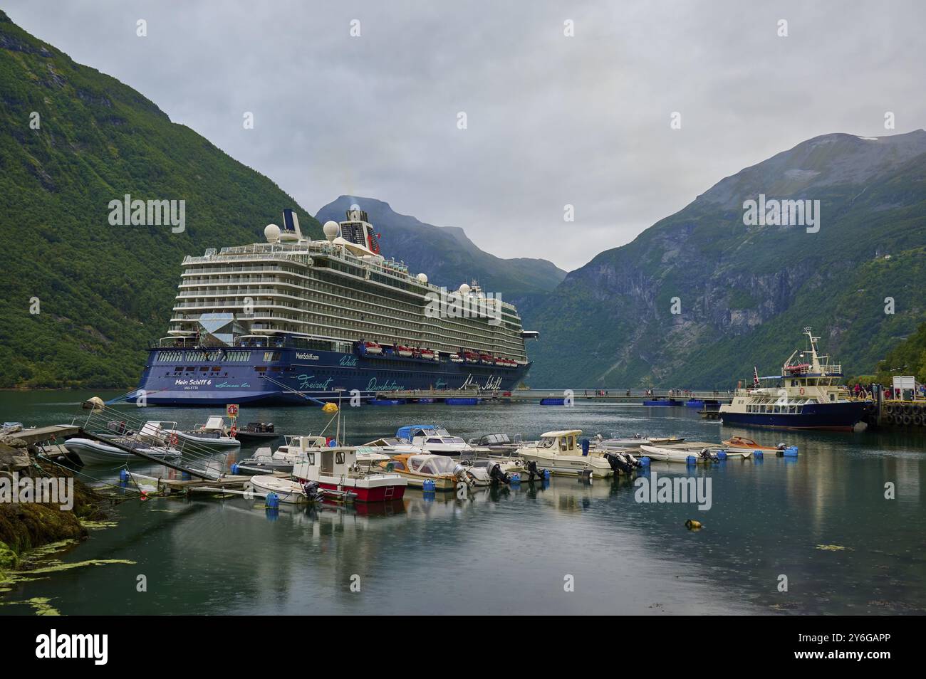 Un grand bateau de croisière, mon bateau 6, se trouve dans le port d'un fjord, entouré de petits bateaux et de montagnes verdoyantes sous un ciel nuageux, Geiranger, Geirang Banque D'Images