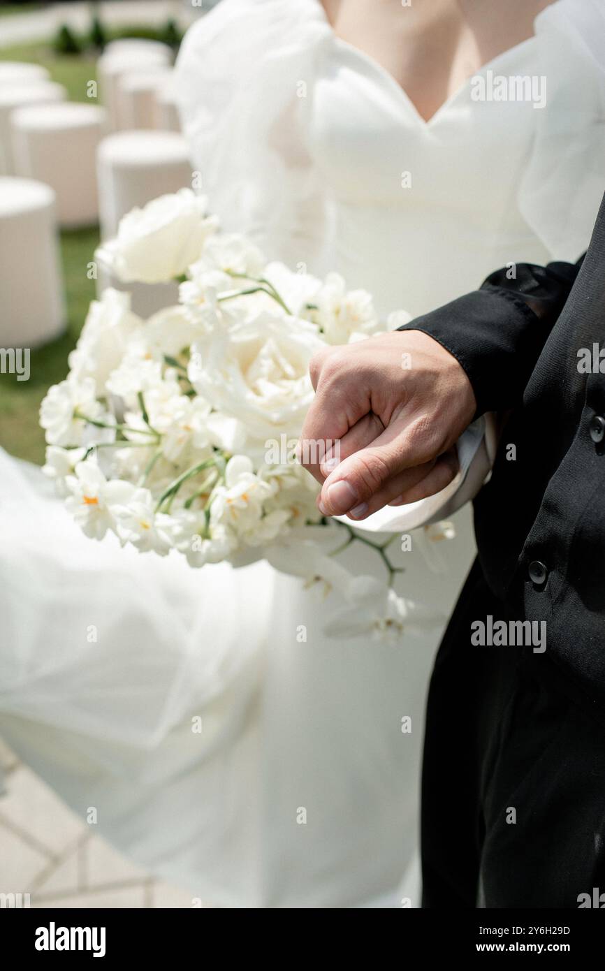 Le marié se tient fièrement, tenant un délicat bouquet de fleurs blanches, ajoutant une touche d'élégance à la joyeuse cérémonie de mariage en plein air sous la bri Banque D'Images