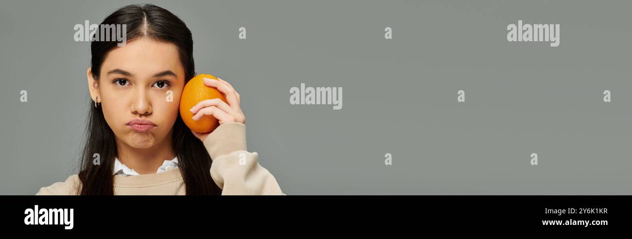 Une jeune femme en tenue à la mode tient une orange sur sa joue, perdue dans la pensée et l'émotion. Banque D'Images