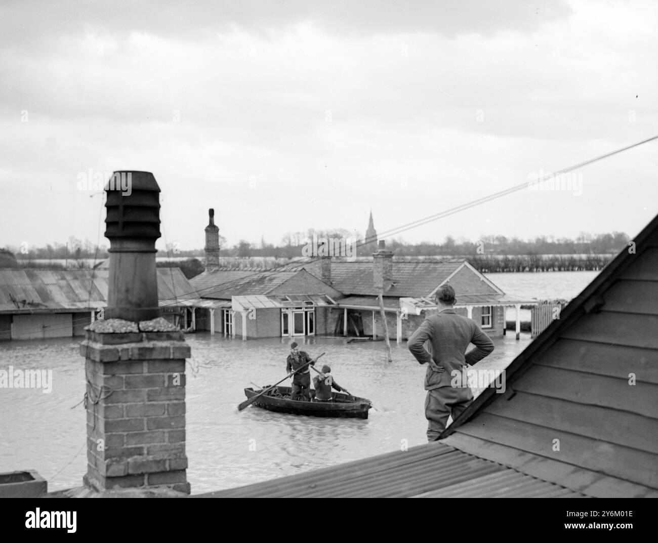 S'accrocher à leur petite tenue. M. J. Harlow attend sur le toit de son bungalow 'Riverside' comme son frère, M. H.V. Harlow et son beau-frère M. D./ Rowlands approche en bateau avec les rations. Qui vivent actuellement dans le grenier d'un de leurs bungalows attenant inondés près de Over, Cambridgeshire, disent : 'le Blitz ne nous a pas chassés de Over' ceci en dépit du fait que leurs maisons et leur petite exploitation, se sont construites en une entreprise florissante depuis que les frères ont déménagé de Londres à Over après que l'un d'eux ait servi dans un aérodrome voisin pendant la guerre. Leurs épouses et enfants Ar Banque D'Images