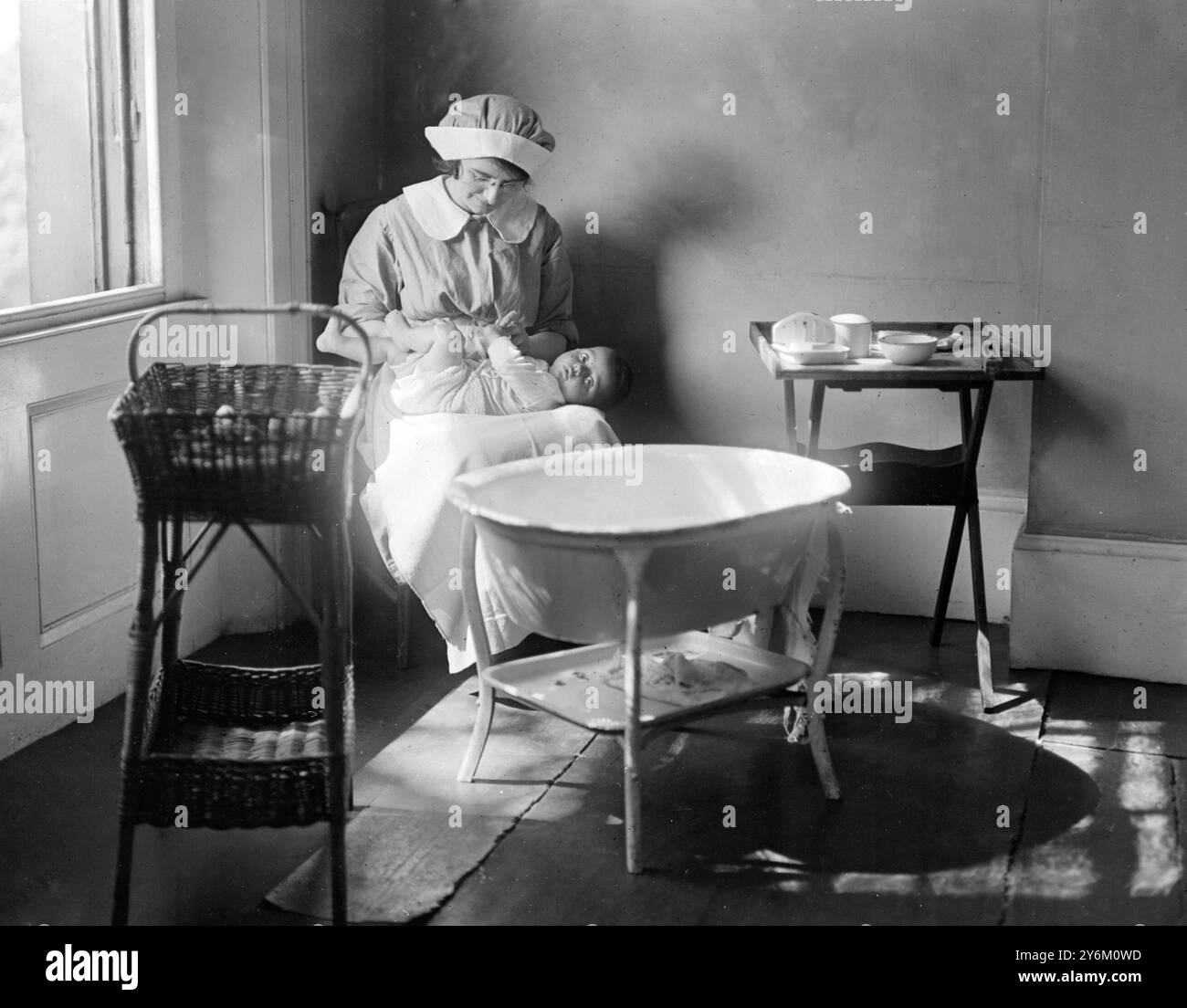 Ordre du bain. Infirmière assise sur une chaise basse, portant mackintosh et tabliers en flanelle, avec bébé sur ses genoux. Le bain est en face d'elle, panier d'un côté et table contenant lotion boracique, coton, etc, de l'autre. Les vêtements de bébé, disposés dans le bon ordre à mettre, et les serviettes turques et faciales devraient être aérées sur le garde-feu à ses côtés. Banque D'Images