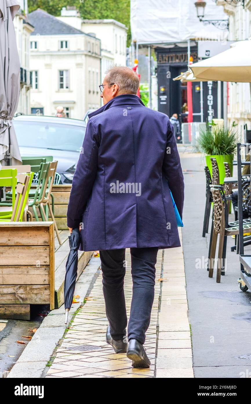 Vue arrière d'un homme portant un imperméable et portant un parapluie marchant sur le trottoir - Tours, Indre-et-Loire (37), France. Banque D'Images