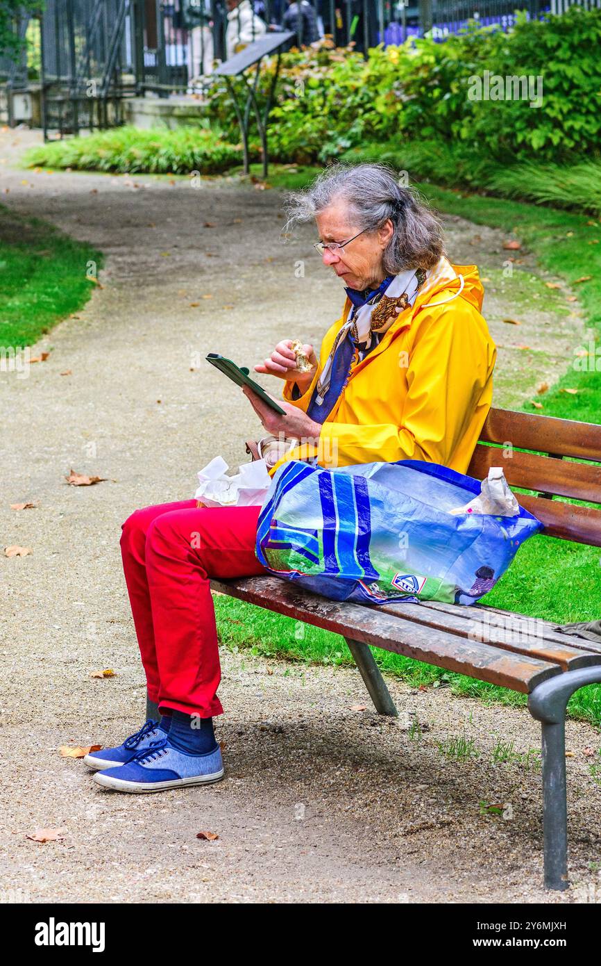 Femme âgée assise sur un banc de parc de centre-ville mangeant et regardant un smartphone - Tours, Indre-et-Loire (37), France. Banque D'Images