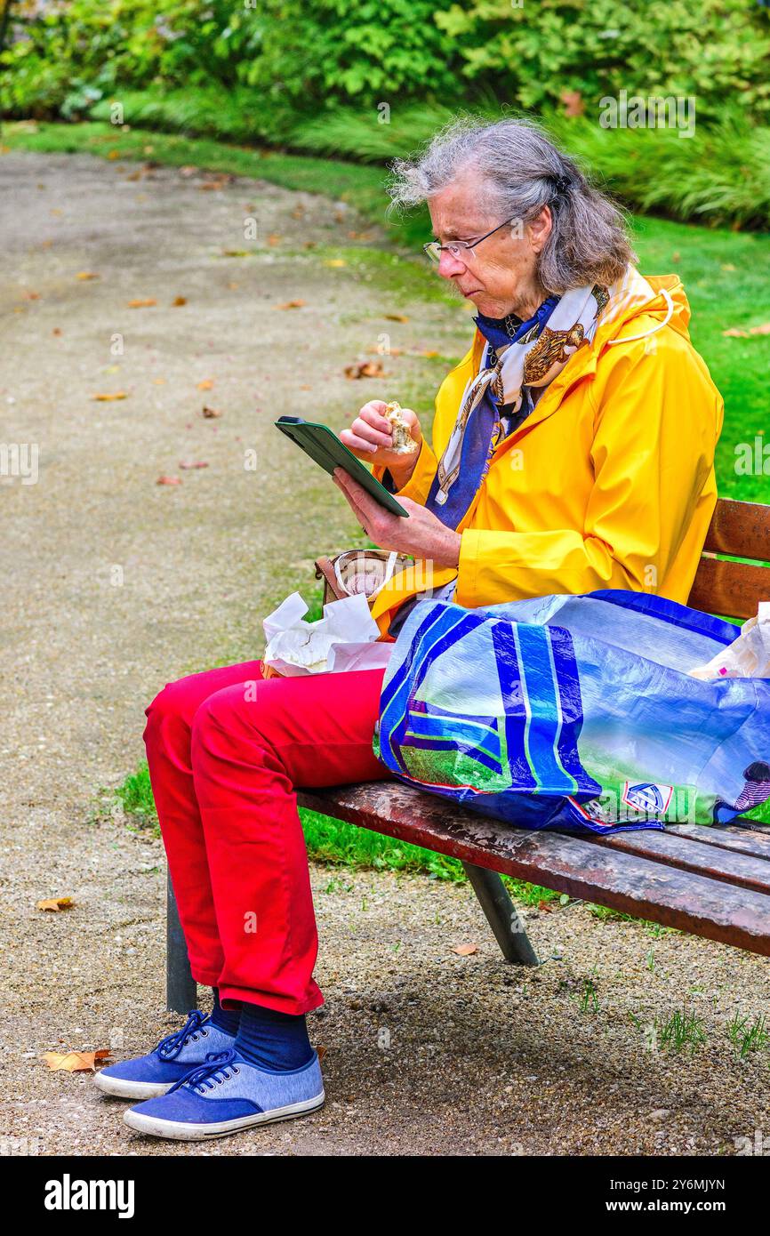 Femme âgée assise sur un banc de parc de centre-ville mangeant et regardant un smartphone - Tours, Indre-et-Loire (37), France. Banque D'Images