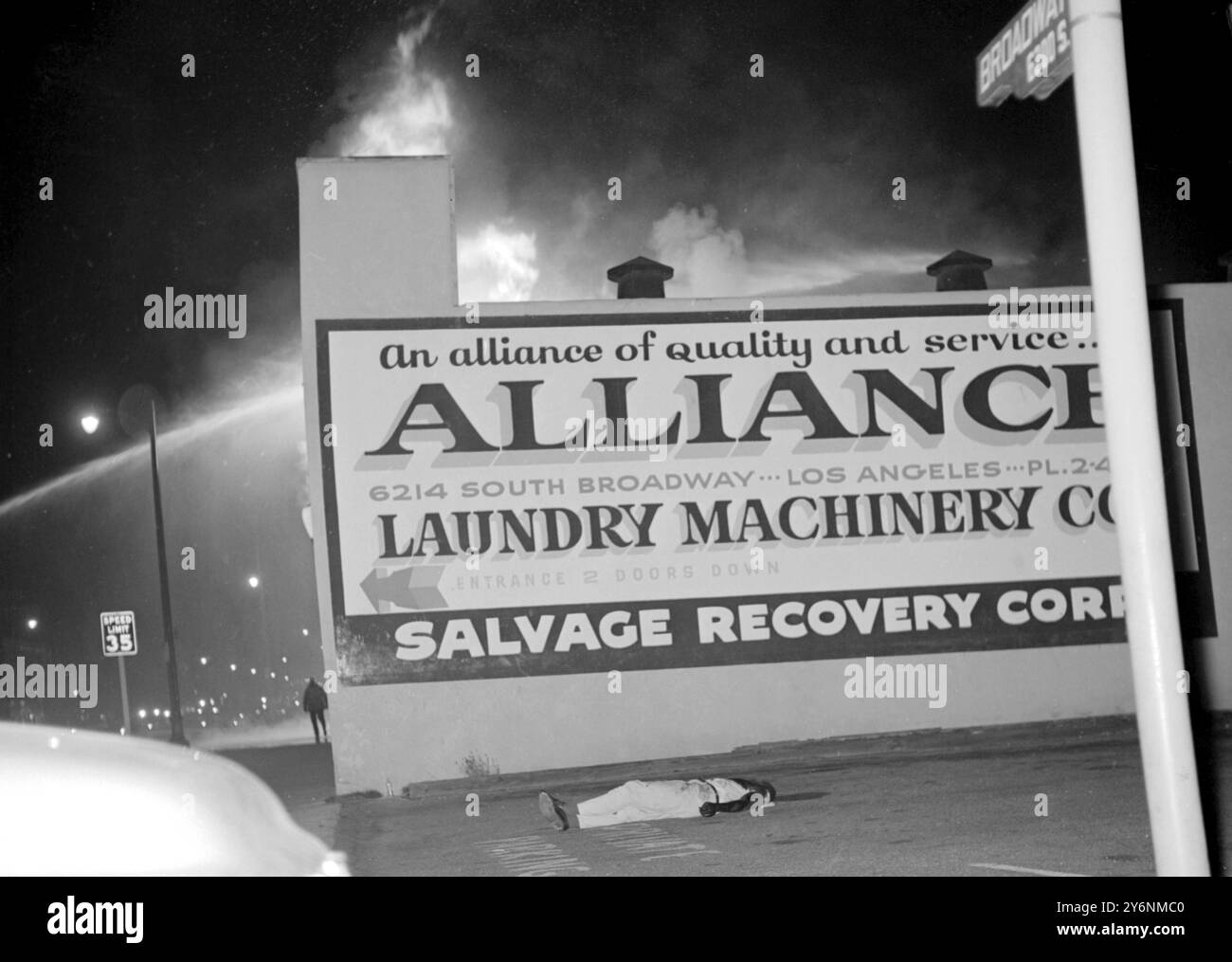 Los Angeles, Californie : le corps d'un pillard mort se trouve à South Broadway ici alors que les bâtiments brûlent derrière le panneau d'affichage Laundry pendant les émeutes elle dans la nuit du 13 au 14 août. Watts district de Los Angeles. Pendant six jours, la zone à prédominance noire a été remplie de violence. - 19 août 1965 Banque D'Images
