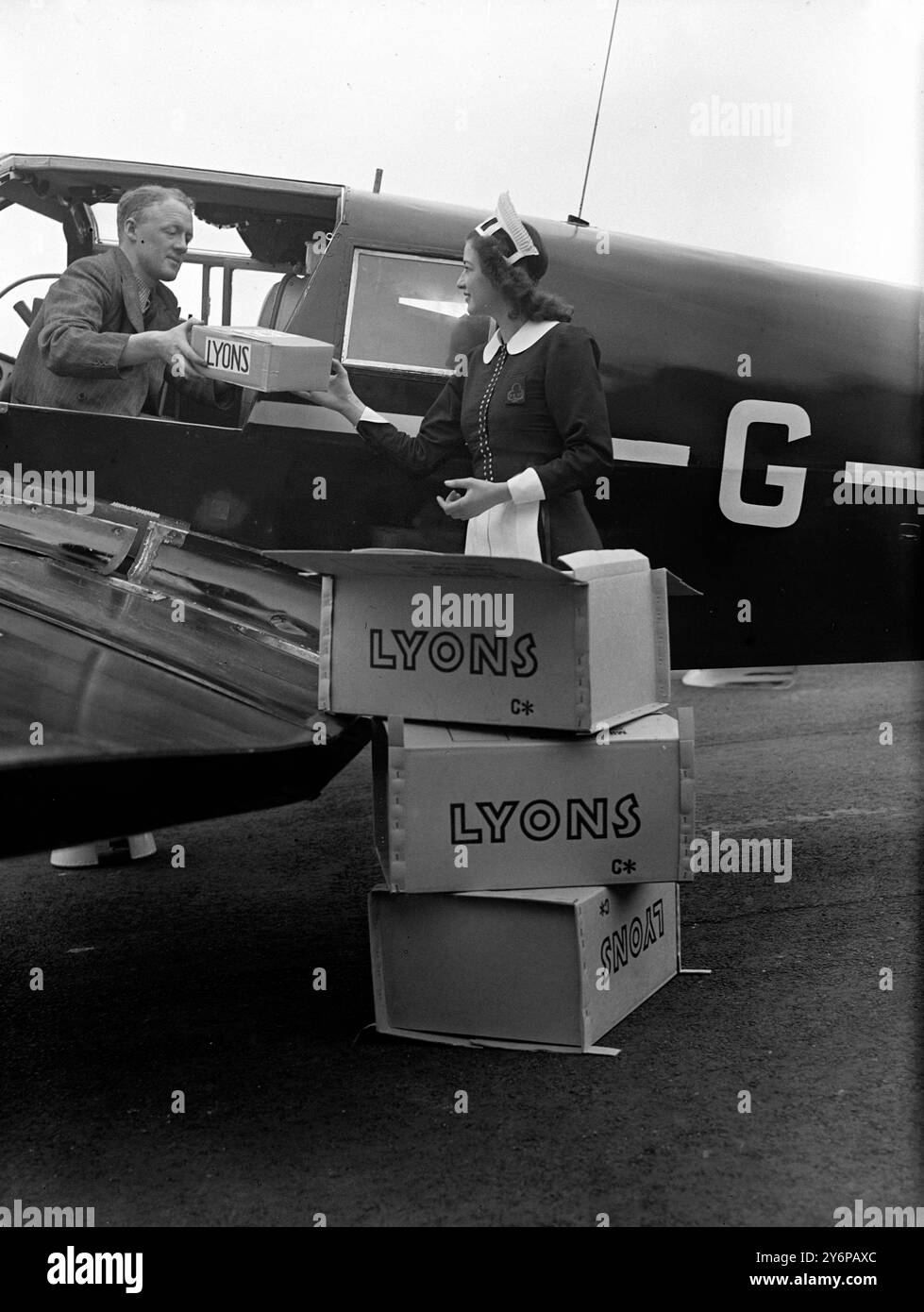 Une serveuse Nippy livrant des marchandises lyonnaises au jeune aviateur néo-zélandais Capt A A Mansfield qui tentera le premier vol record officiel chronométré autour du monde lorsqu'il décollera de l'aéroport de Blackbushe le 21 avril 1948. Le capitaine Mansfield, qui a choisi un avion léger monomoteur Percival Proctor pour sa tentative de record, attaquera également quatre records point à point , Londres - Montréal , Londres - New York , Tokyo - Paris et Karachi - Londres. 19 avril 1948 Banque D'Images