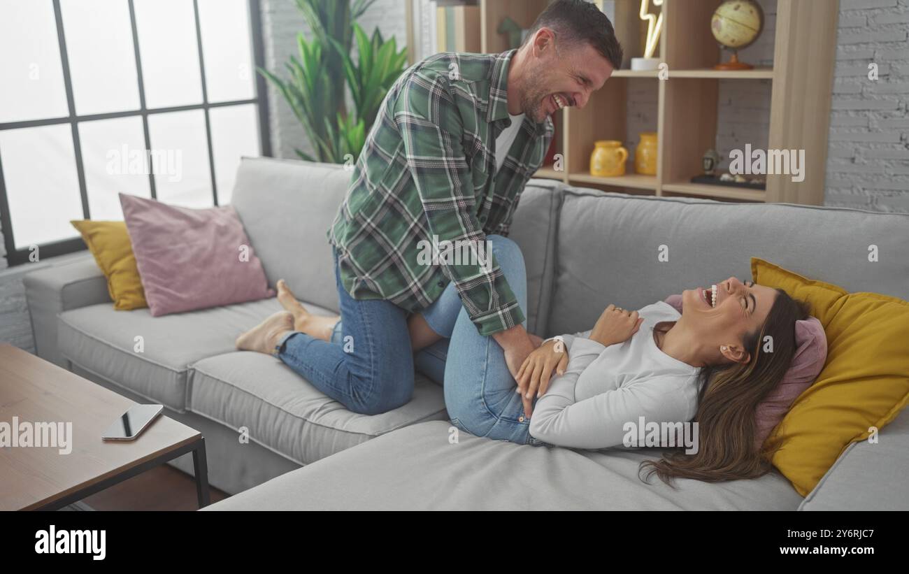 Femme riante chatouillée par un homme joyeux sur un canapé confortable à l'intérieur, mettant en valeur leur relation joyeuse. Banque D'Images