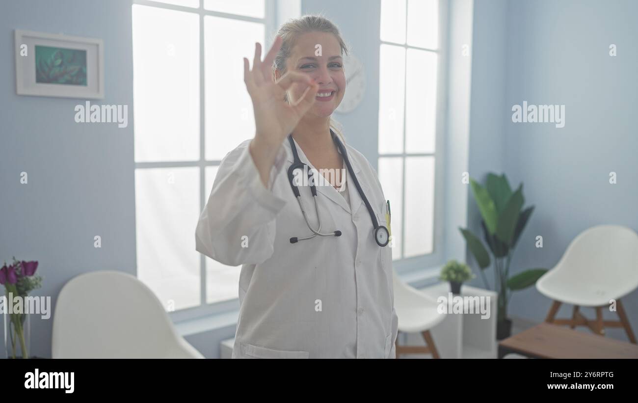 Femme blonde médecin donnant le signe OK dans la salle de clinique lumineuse avec stéthoscope et plantes Banque D'Images