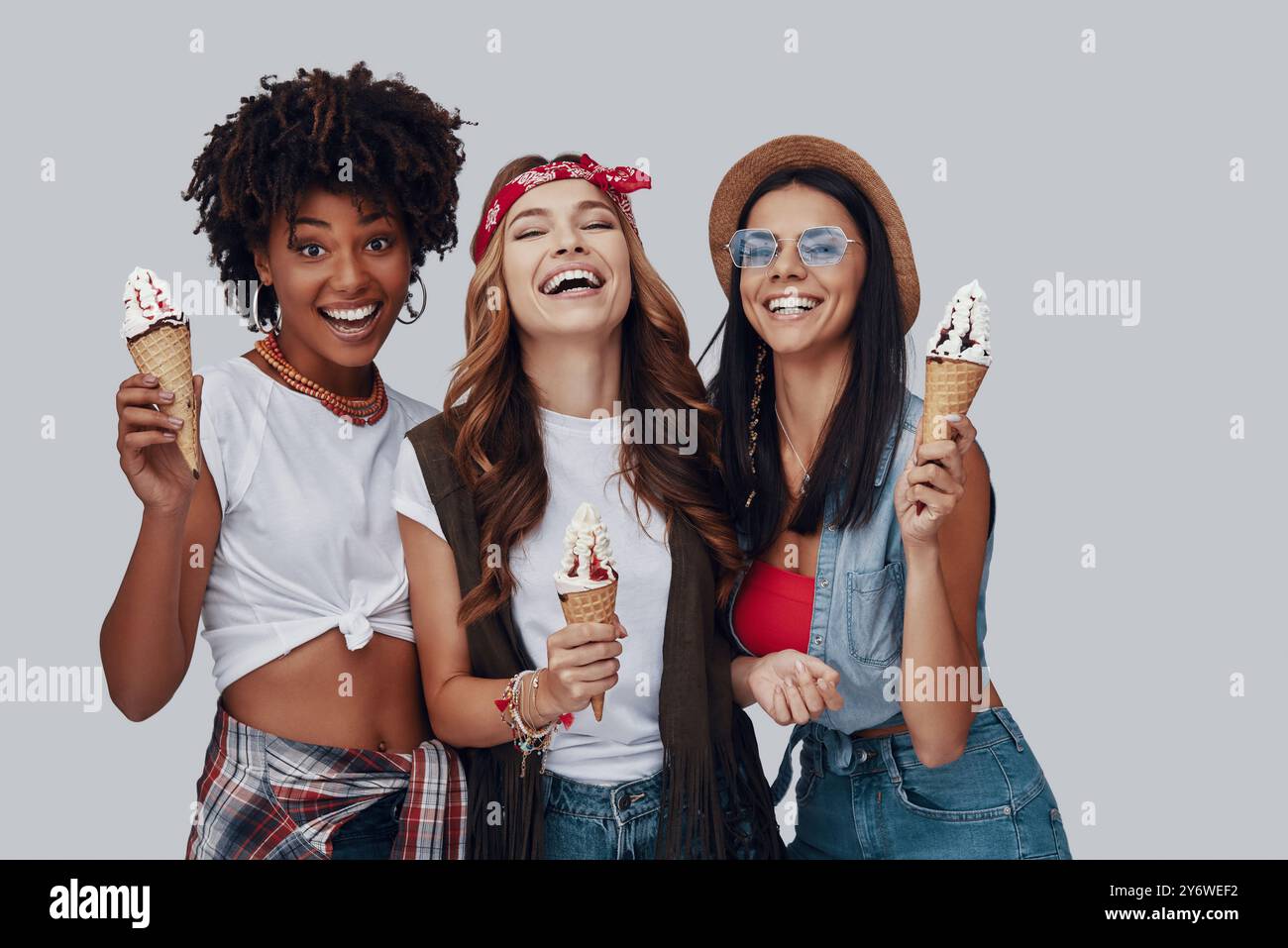 Trois jeunes femmes attrayantes mangeant de la crème glacée et souriant tout en se tenant debout sur fond gris Banque D'Images