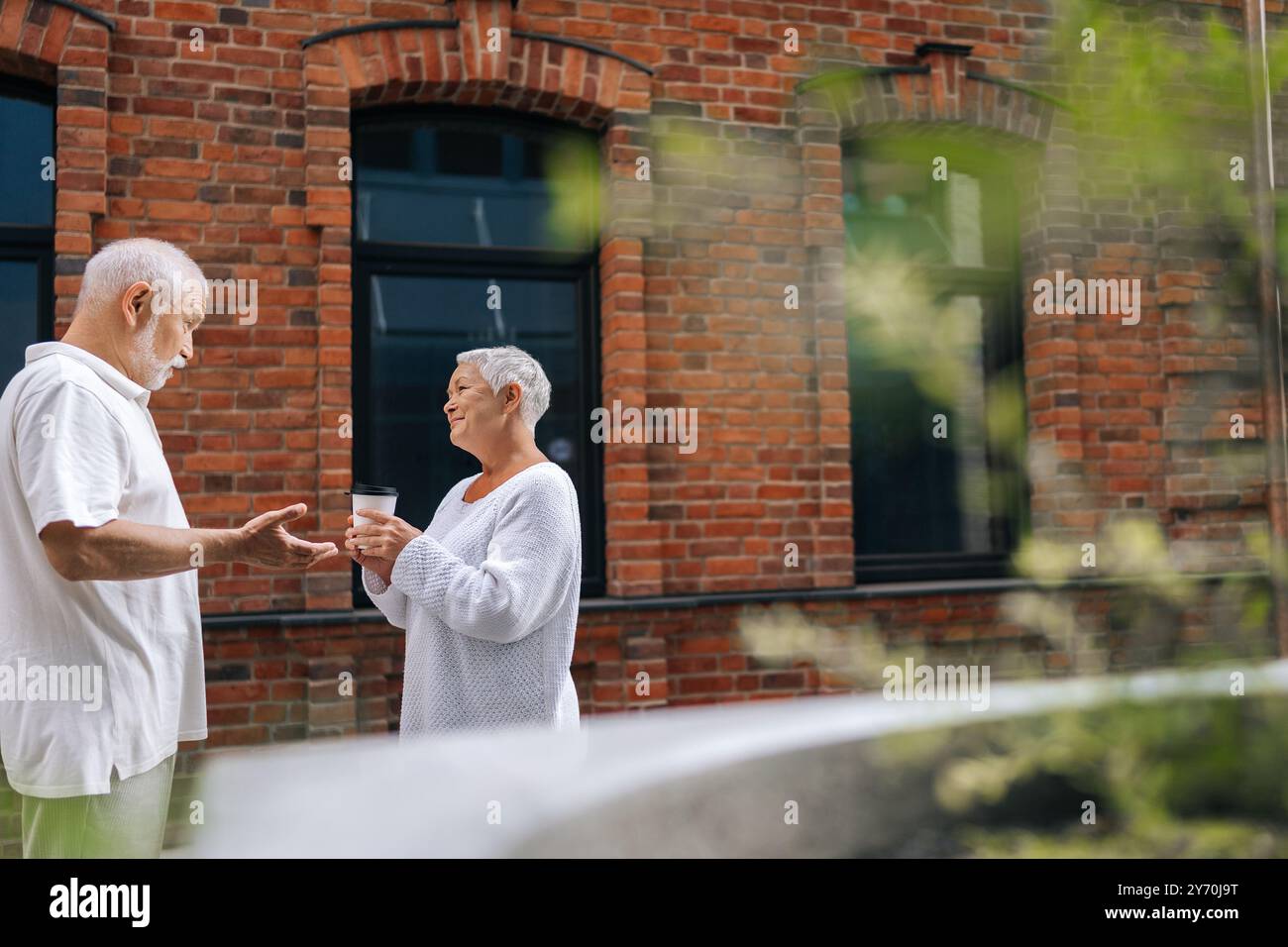 Vue latérale d'un couple âgé discutant joyeusement dans la rue urbaine avec des tasses à café, savourant la chaude journée d'été. Bel homme senior et femme souriant Banque D'Images