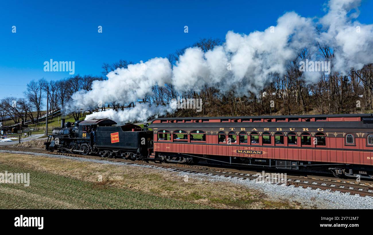 Ronks, Pennsylvanie, États-Unis, 18 février 2024 - Une locomotive à vapeur d'époque se déplace le long d'un chemin de fer panoramique, soufflant de la fumée blanche contre un ciel bleu vif. Le train traverse des paysages verdoyants, mettant en valeur son charme nostalgique. Banque D'Images