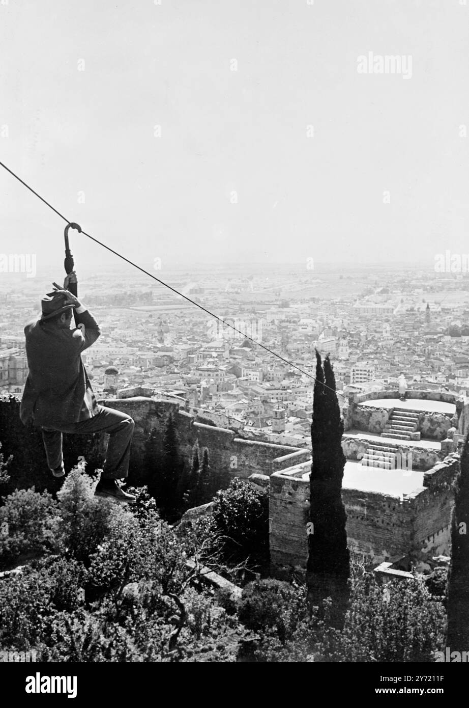 Rouler sur un Brolly . Malaga ; Espagne : L'acteur britannique Denholm Elliot tient son chapeau alors qu'il commence une descente précaire de 350 pieds de câble d'acier, d'une tour de transmission de Radio Nacional de Espana - en utilisant son parapluie comme ' télésiège ' . Ci-dessous , l'héroïne de la production de Michael Todd Junior ' Scent of Mystery ' se trouve sur l'ancienne forteresse maure , Gilbralfaro , ignorant qu'Elliot court pour la rejoindre avant le méchant . Cette scène passionnante atteint le point culminant du film. Pendant la fusillade , trois médecins , une infirmière et deux ambulances sont restés là , en cas d'accident . Denholm Ell Banque D'Images