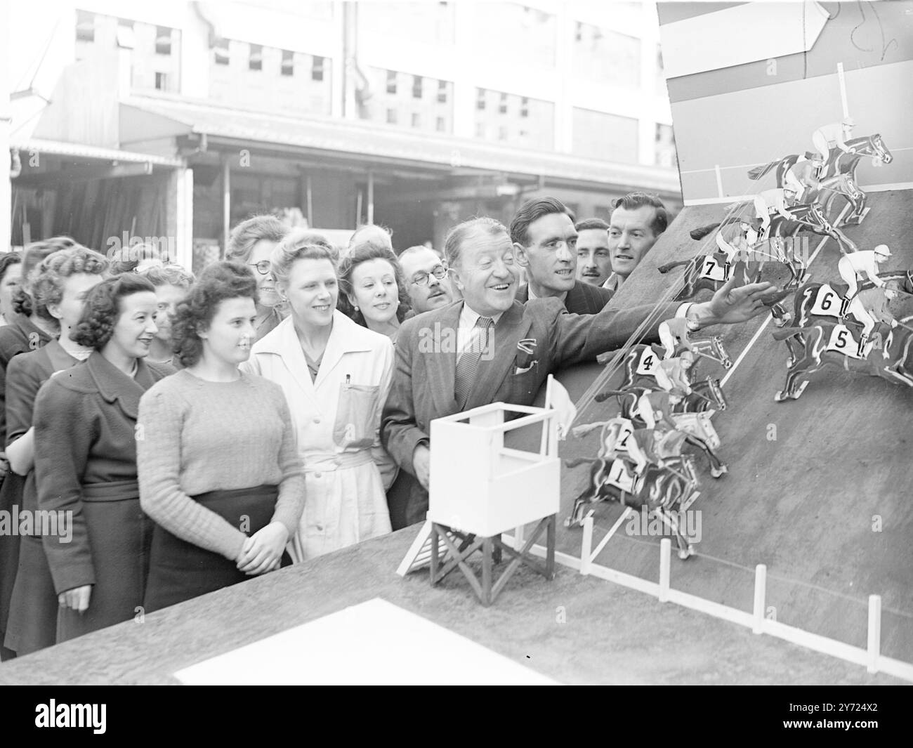 Tommy Handley était sur place à l'usine Hoover, Great West Road, aujourd'hui (vendredi) pour donner un coup d'envoi aux enjeux « anti-Waste » - une idée originale selon laquelle les ouvriers de l'usine rivalisent en équipes pour réduire les déchets et aider ainsi la production. La firme espère que ce système permettra d'économiser environ 75 000 £ par an. 10 chevaux réalistes représentant chacun une équipe se déplaceront le long d'un « hippodrome » pour marquer la réalisation des économies de déchets. Images : Tommy Handley, avec une foule de travailleurs heureux, donne sa bénédiction aux enjeux «anti-Waste» à l'usine Hoover, aujourd'hui (vendredi). 16 avril 1948. Banque D'Images