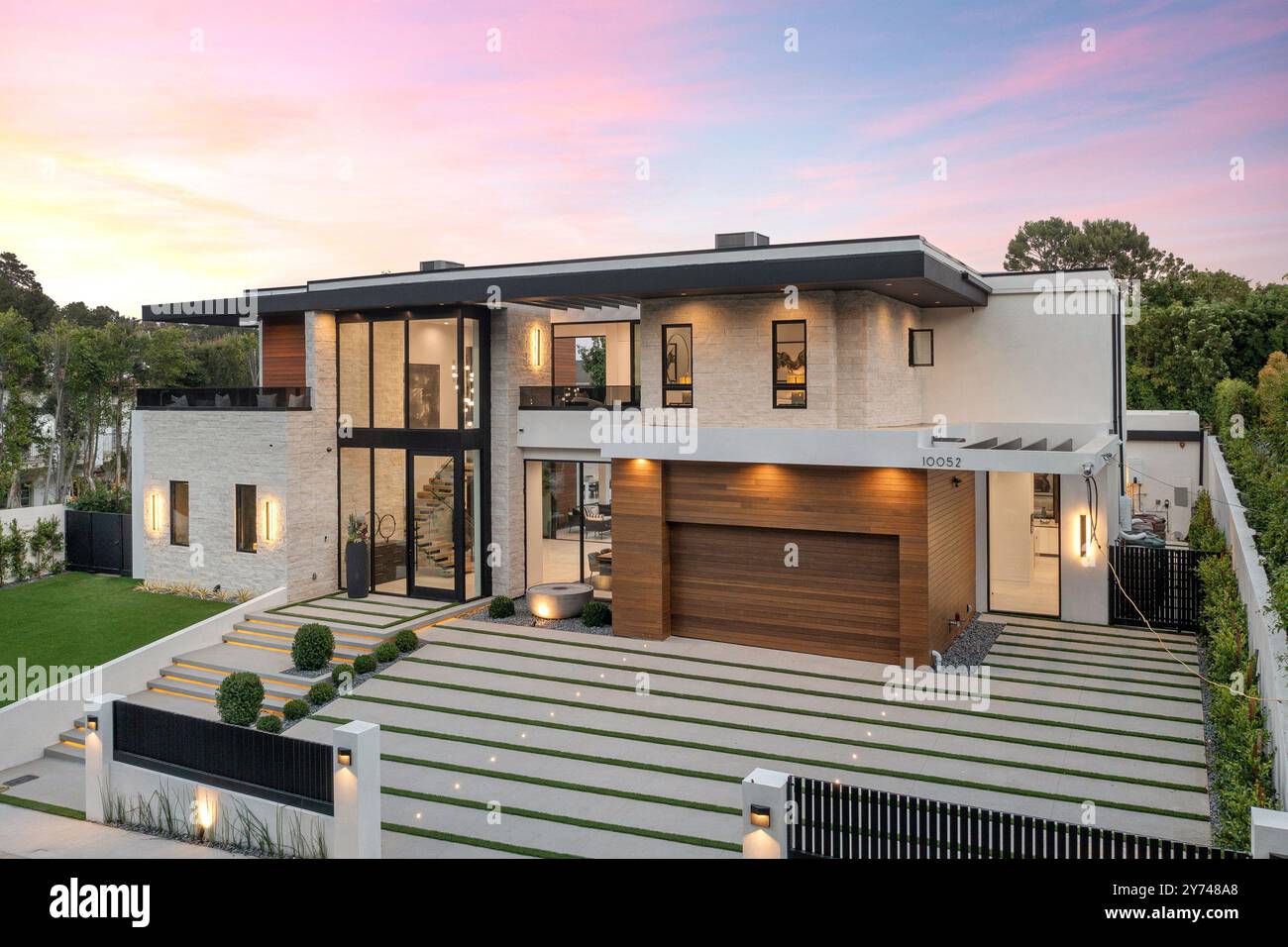 Extérieur de la maison de luxe moderne avec de grandes fenêtres, porte de garage en bois et pelouse bien entretenue au coucher du soleil. Banque D'Images