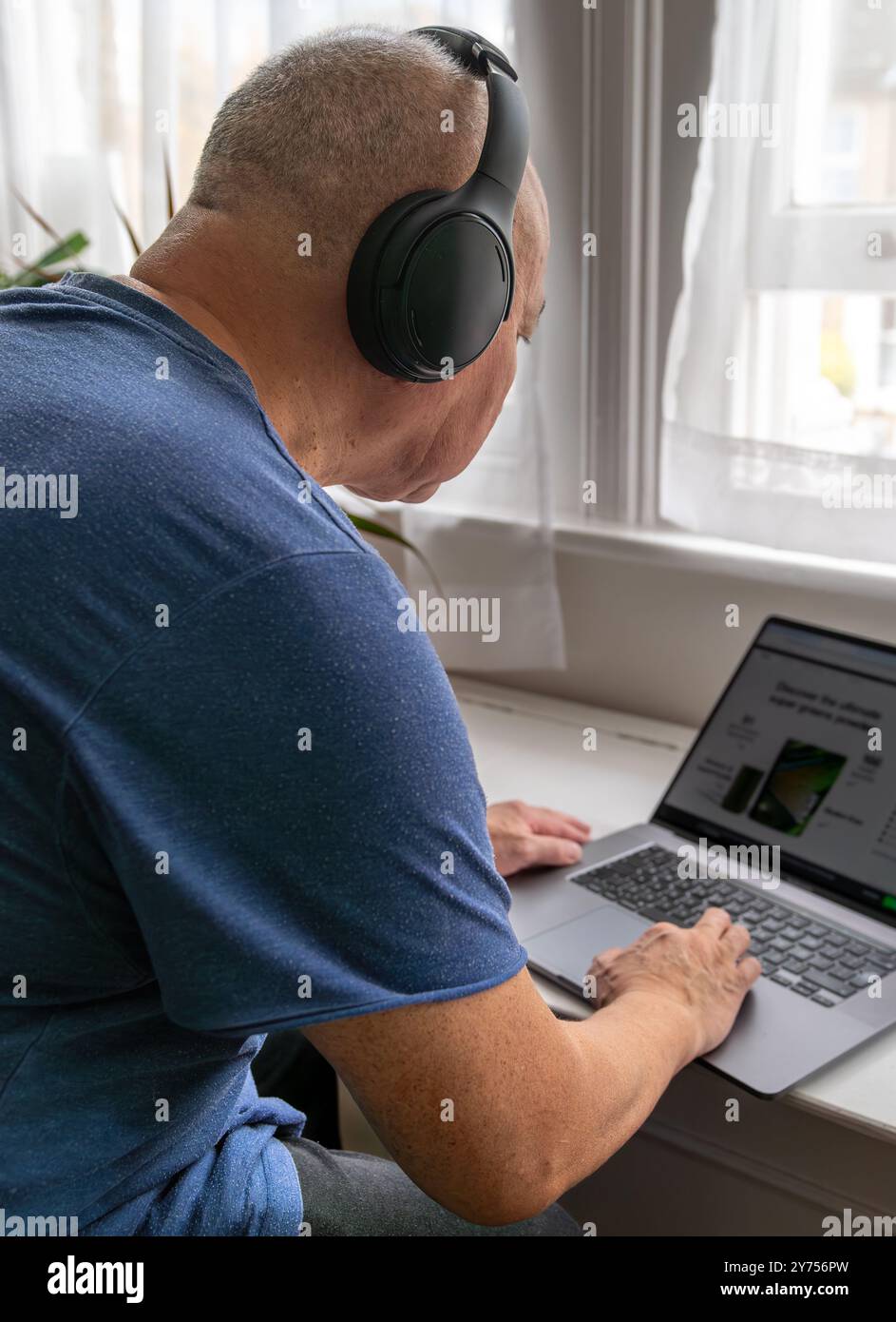 Un homme âgé à la maison avec un casque sans fil et un ordinateur portable, apprenant et essayant de se tenir à jour avec la technologie moderne. Une utilisation technologique contre Banque D'Images