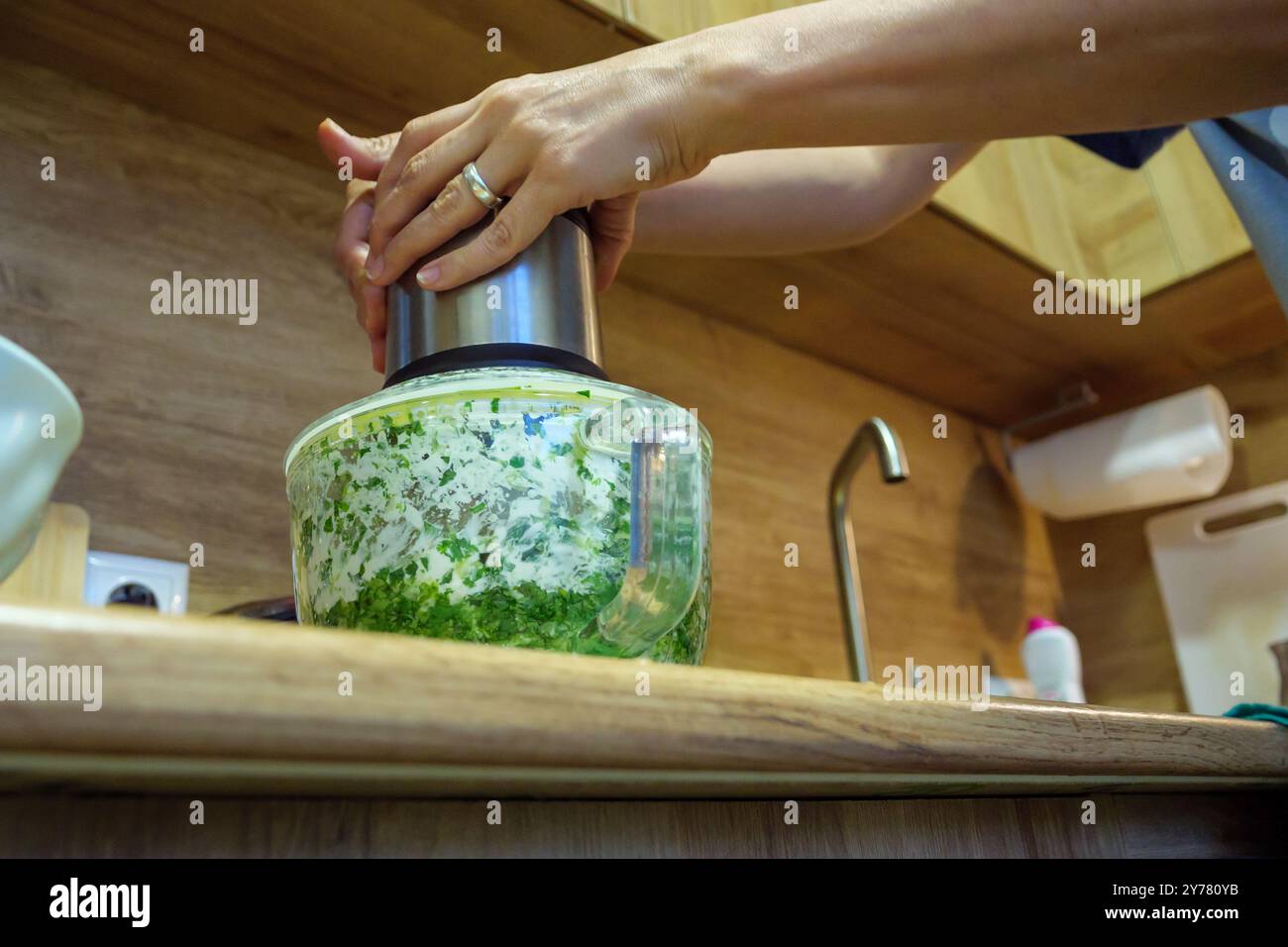 Les mains pressent vers le bas un couvercle de broyeur de nourriture rempli d'herbes hachées et d'ingrédients sur un comptoir en bois dans une cuisine moderne, pendant la préparation des repas. Banque D'Images