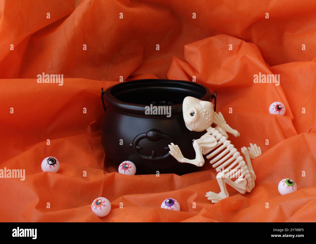 Squelette de grenouille mort effrayant essayant d'entrer dans un chaudron noir de sorcière vide. Décoration amusante de vacances d'Halloween avec fond de tissu de couleur orange. Banque D'Images