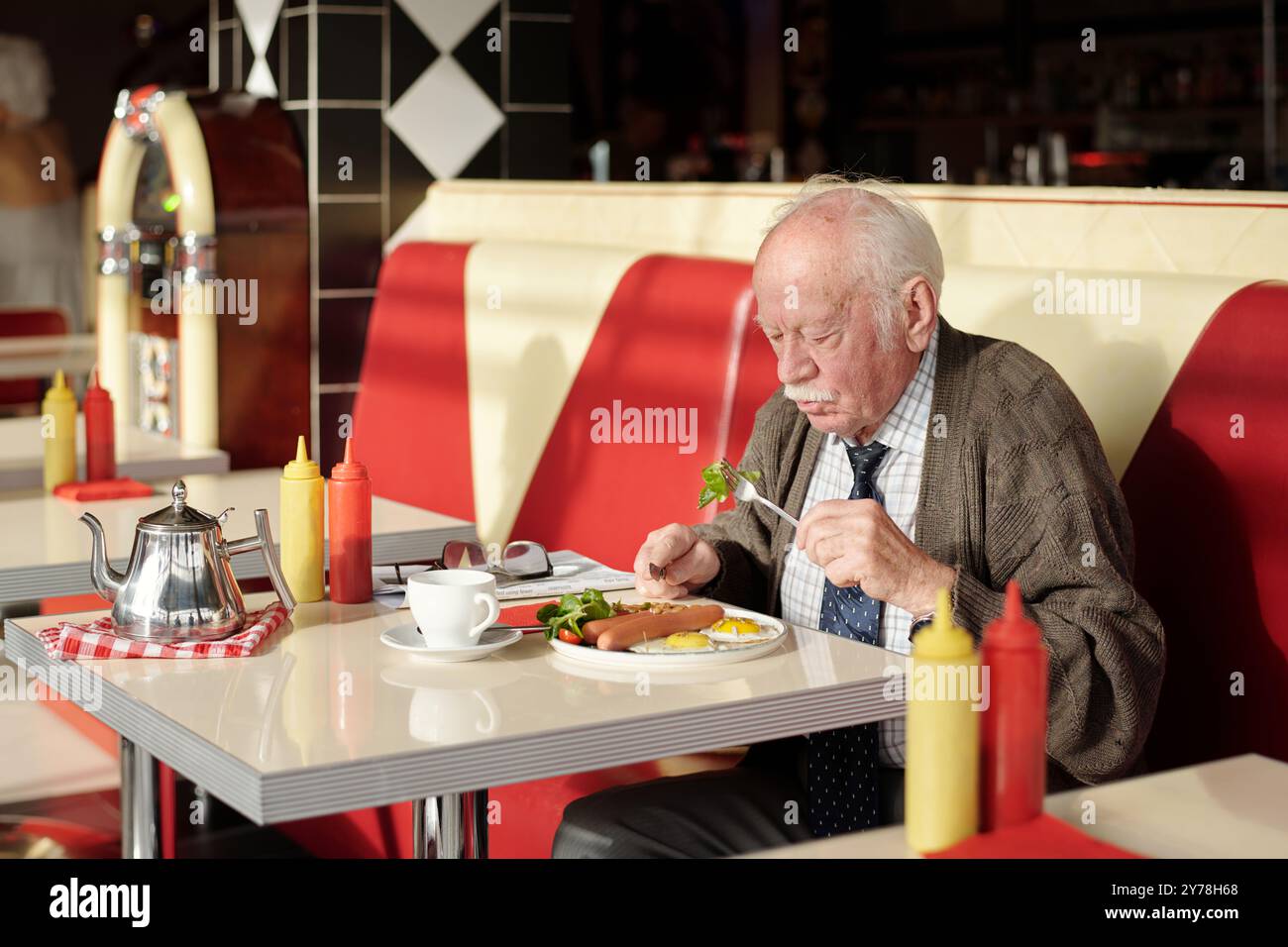 Senior dégustant un copieux petit-déjeuner dans un légendaire restaurant américain rétro avec cabines rouges, juke-box vintage et condiments sur table créant une atmosphère nostalgique rappelant les années 1950 Banque D'Images