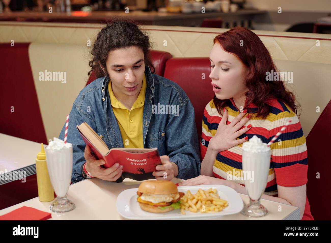 Deux personnes assises dans une cabine de diner, engagées dans une conversation tout en tenant un livre et divers repas sont présents. La personne aux cheveux roux semble surprise, les milkshakes et la nourriture sur la table Banque D'Images