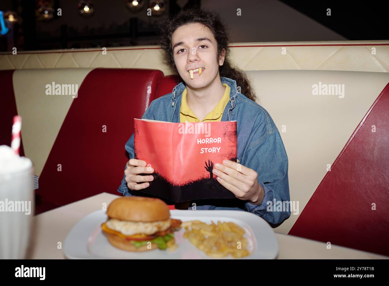 Individu en chemise bleue tenant livre d'histoire d'horreur tout en étant assis dans la cabine de restaurant avec hamburger et frites sur la table et paille à distance Banque D'Images