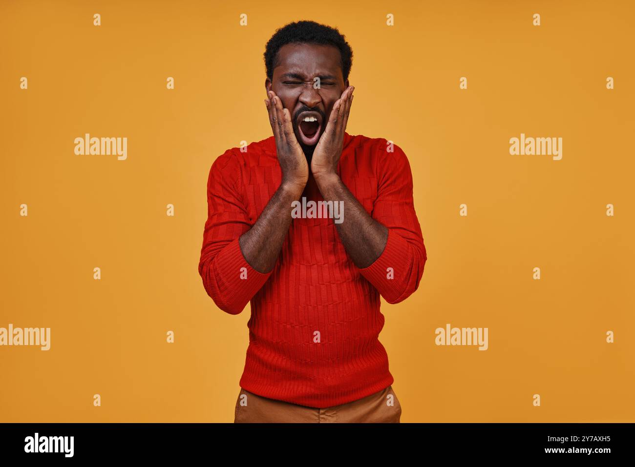 Frustré jeune homme africain dans des vêtements décontractés tenant le visage dans les mains tout en se tenant debout sur fond jaune Banque D'Images