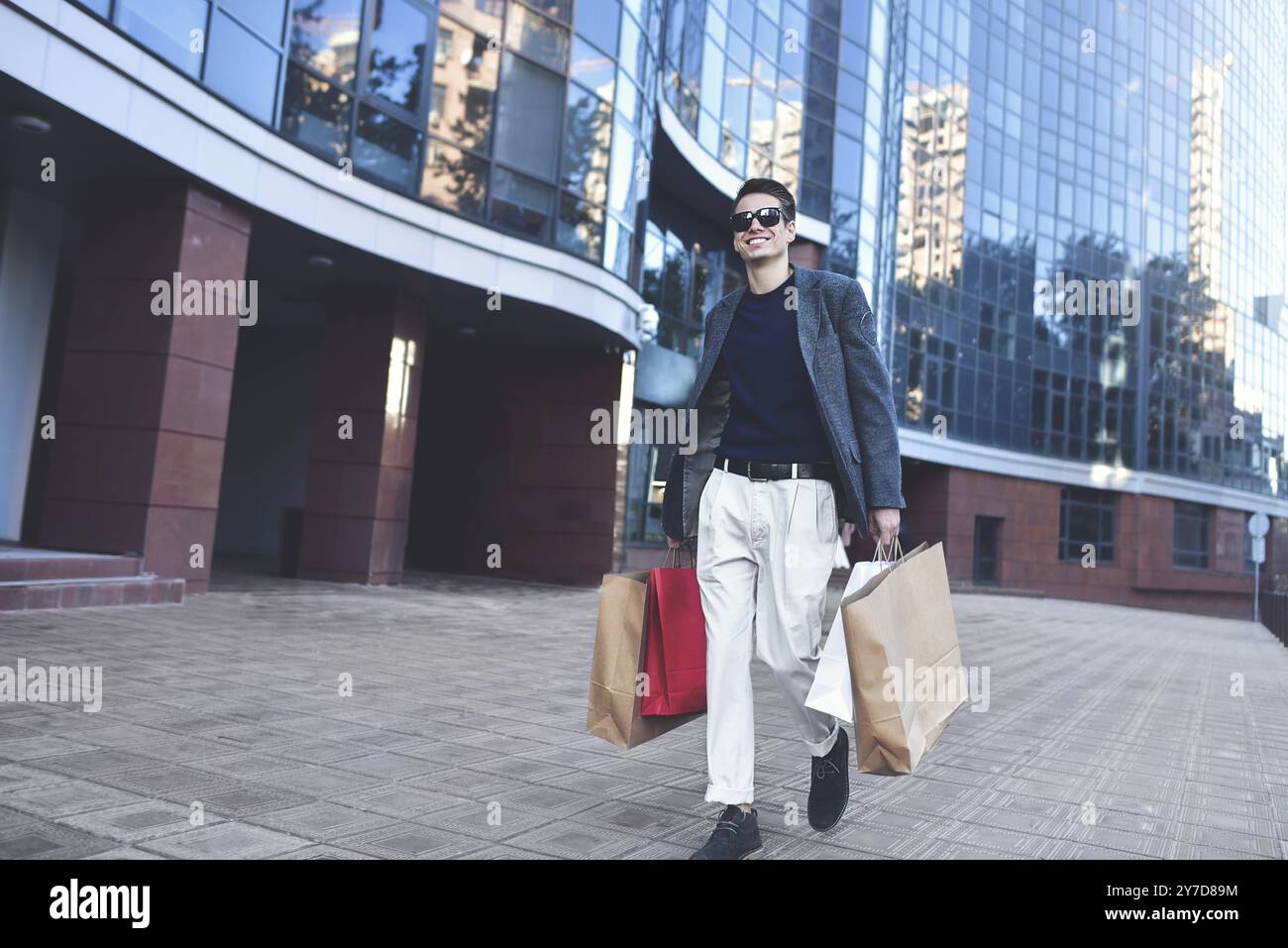 Positif gars cool avec des lunettes de soleil et des sacs en papier marchant près de la vitrine avec réflexion Banque D'Images