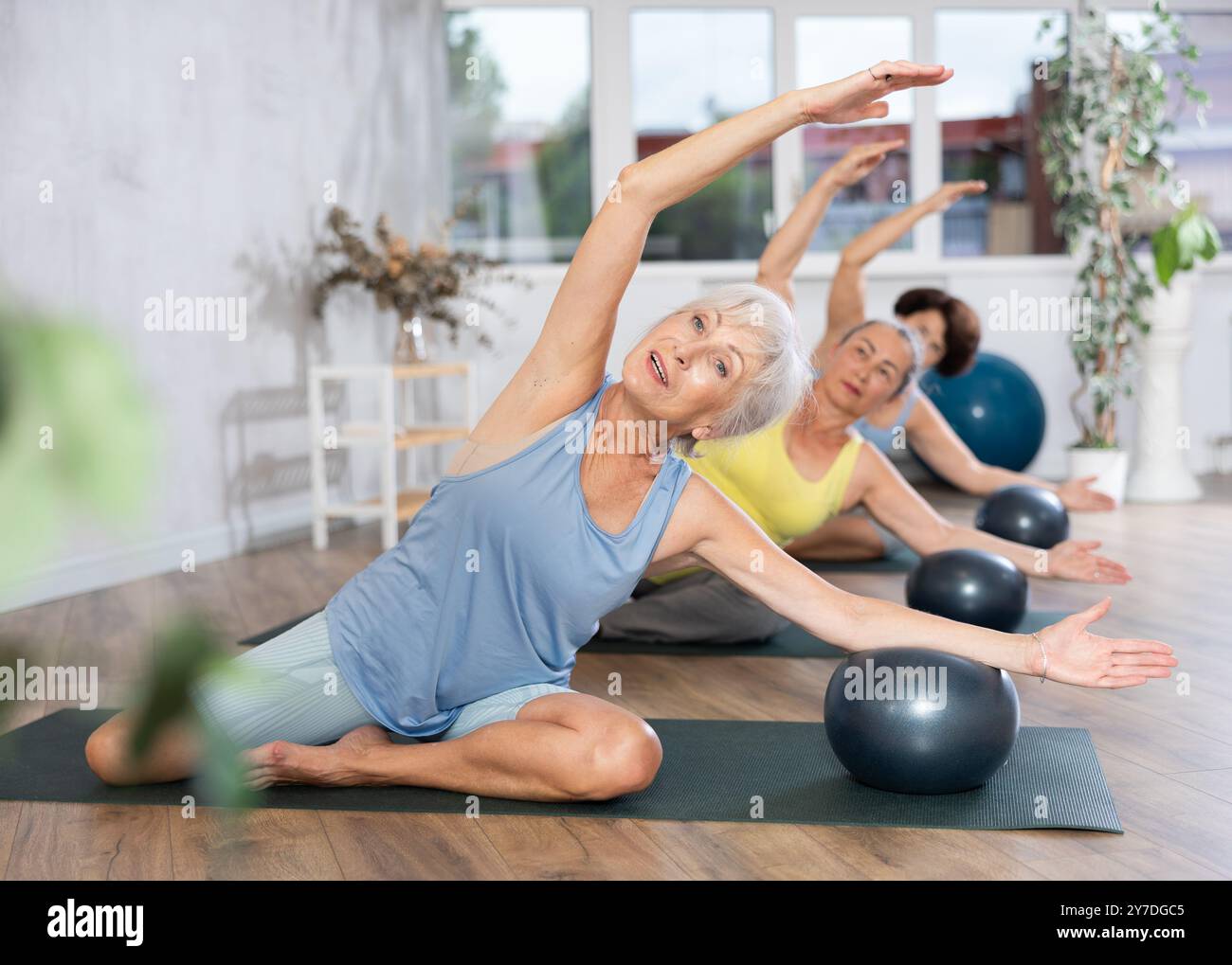 Vieille femme pratiquant la pose demi-lotus avec softball sous le bras dans la salle de fitness légère Banque D'Images