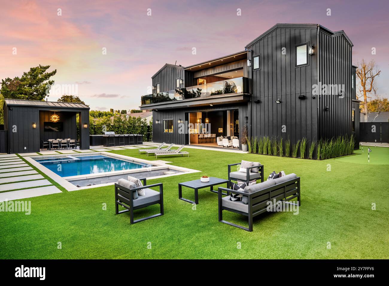 Une maison noire moderne avec une cour arrière luxueuse avec une piscine, des sièges en plein air et une pelouse bien entretenue au coucher du soleil. Banque D'Images