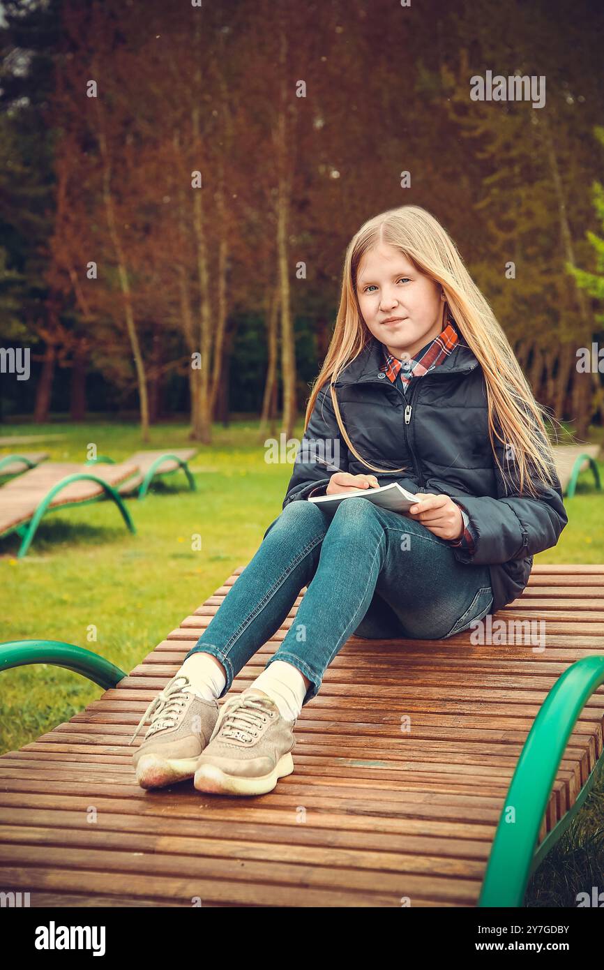 Une fille est assise sur un banc extérieur avec un cahier, entourée d'arbres et d'herbe par une journée ensoleillée Banque D'Images