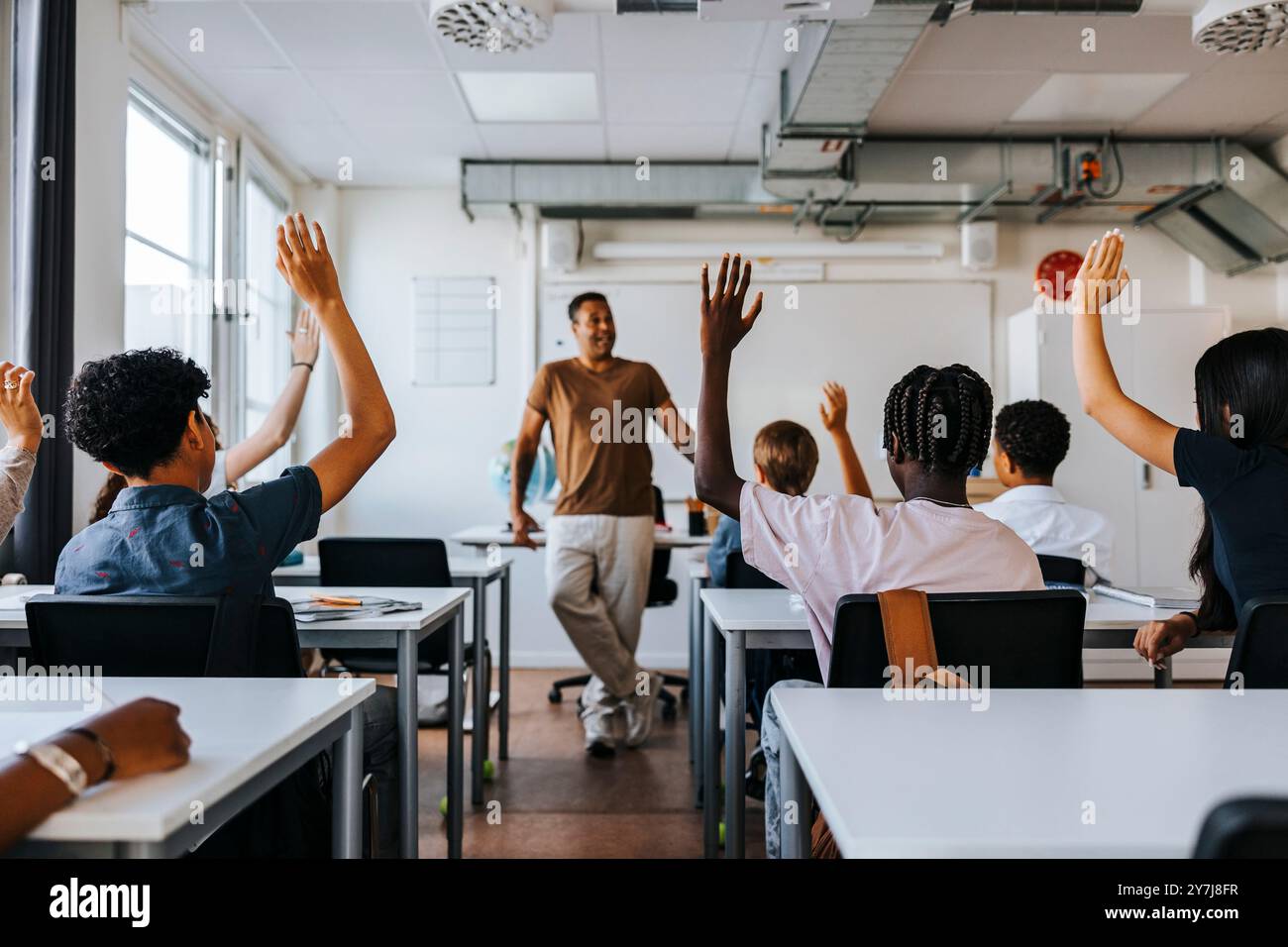 Vue arrière des élèves du premier cycle du secondaire masculin et féminin avec la main levée alors que l'enseignant enseigne en classe à l'école Banque D'Images